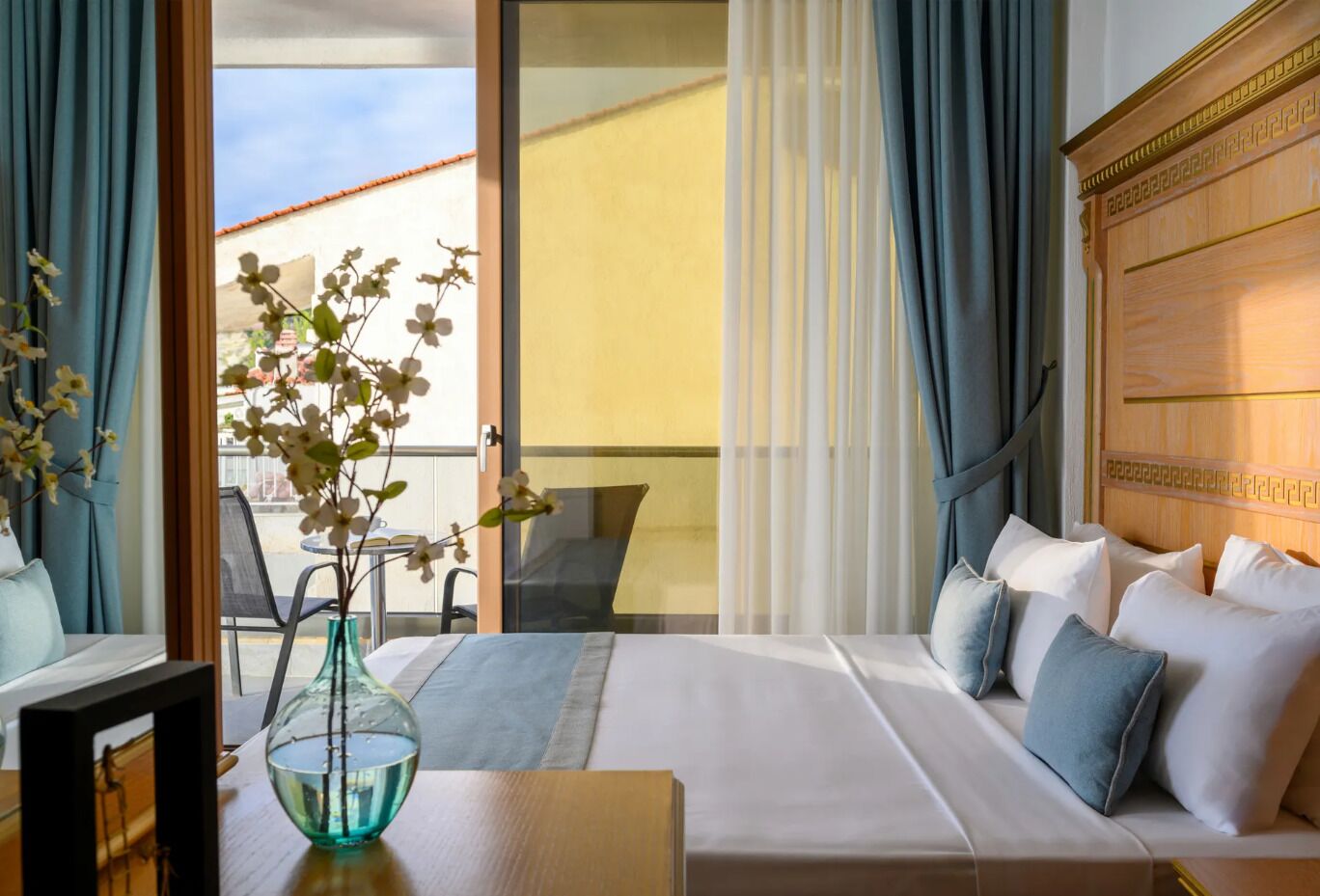 A hotel room with a bed, a vase with flowers, a table, and a view of the outside.