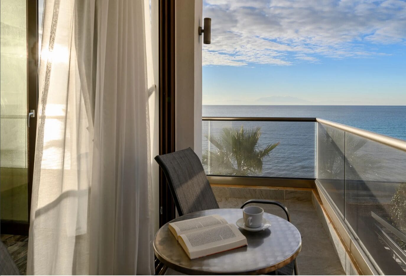 Un balcon donnant sur la mer, une chaise, une table avec un livre et une tasse, et un rideau à moitié tiré.