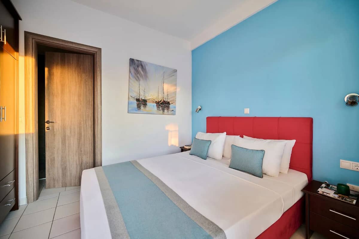 A hotel room with a large bed, a red headboard, a painting of boats on the wall, and a blue accent wall.