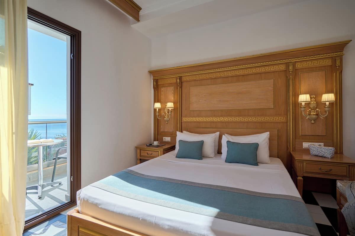 A hotel room with a large bed, two bedside tables with lamps, a view of the sea through a sliding door, and a balcony with a table and chairs.