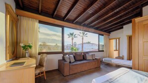 Une chambre d’hôtel avec un plafond en bois, un canapé, un lit, un bureau et une grande fenêtre donnant sur des palmiers et des maisons.