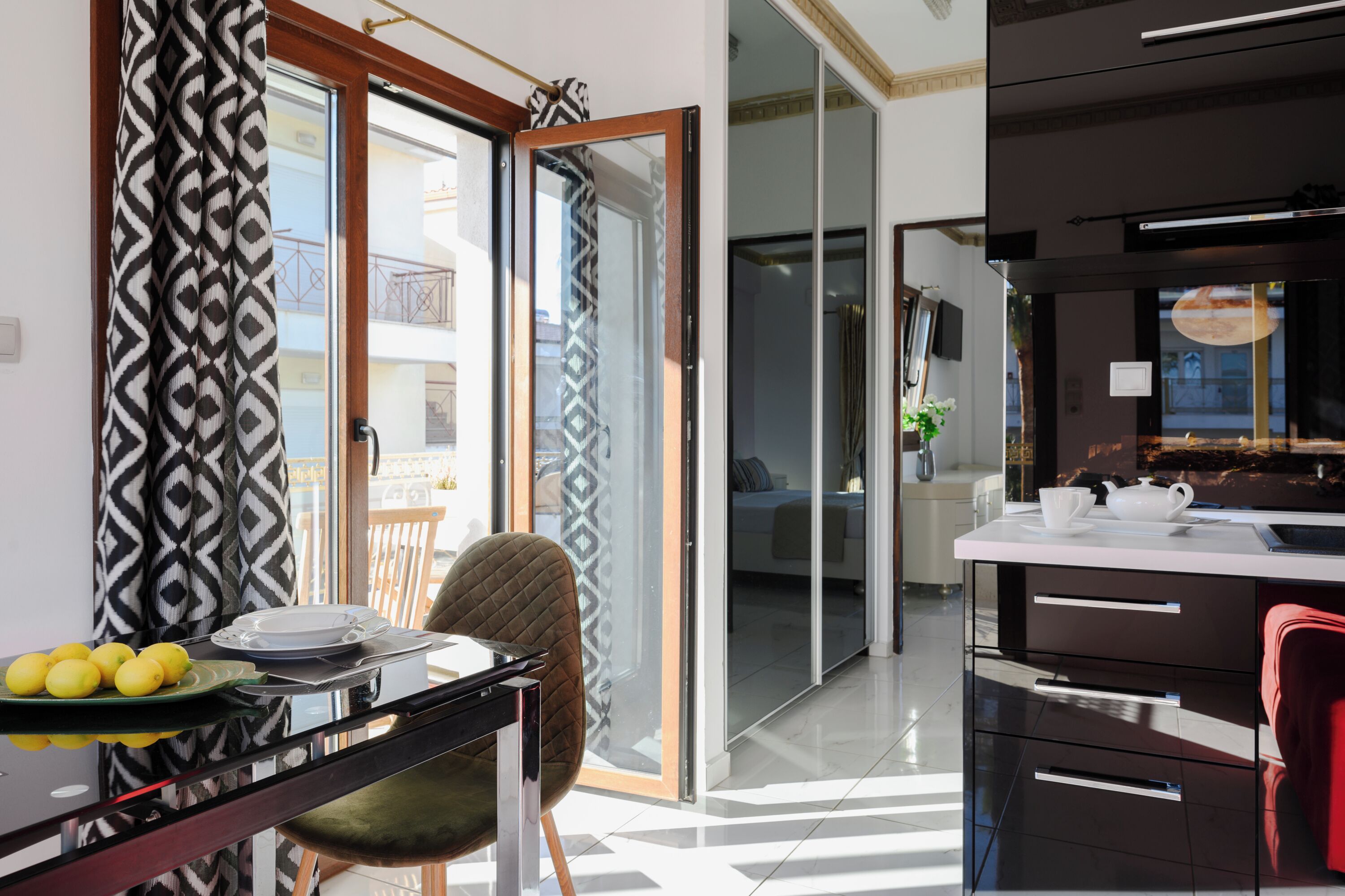 A modern kitchen with a black countertop, a dining area with a glass table, and a view of a balcony with patterned curtains.