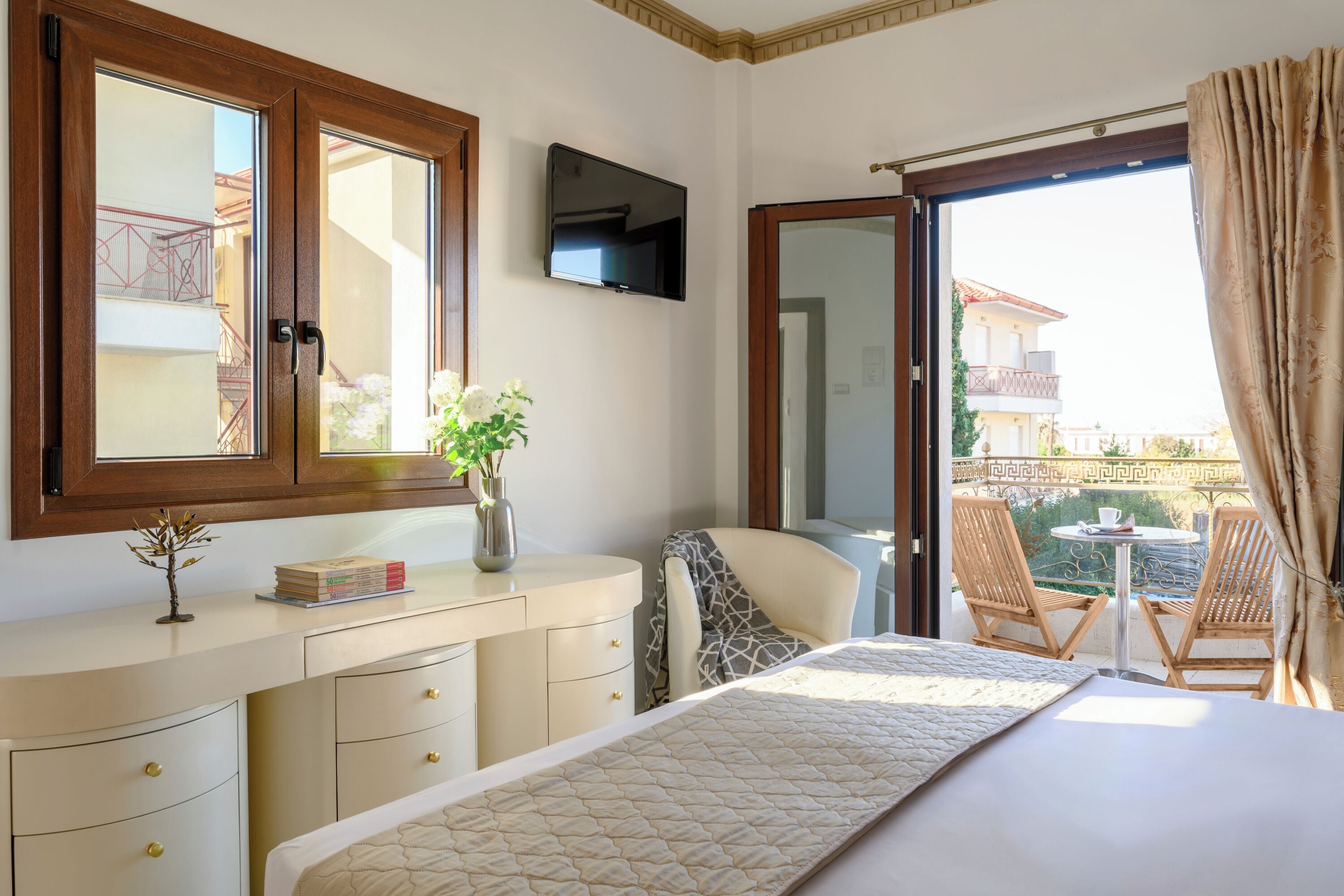 A bedroom with a bed, a TV, a desk with a vase, and a balcony with chairs.