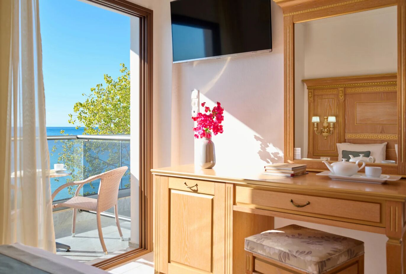 A room with a wooden desk, a television mounted on the wall, a vase with flowers, and a view of the sea through a large window.