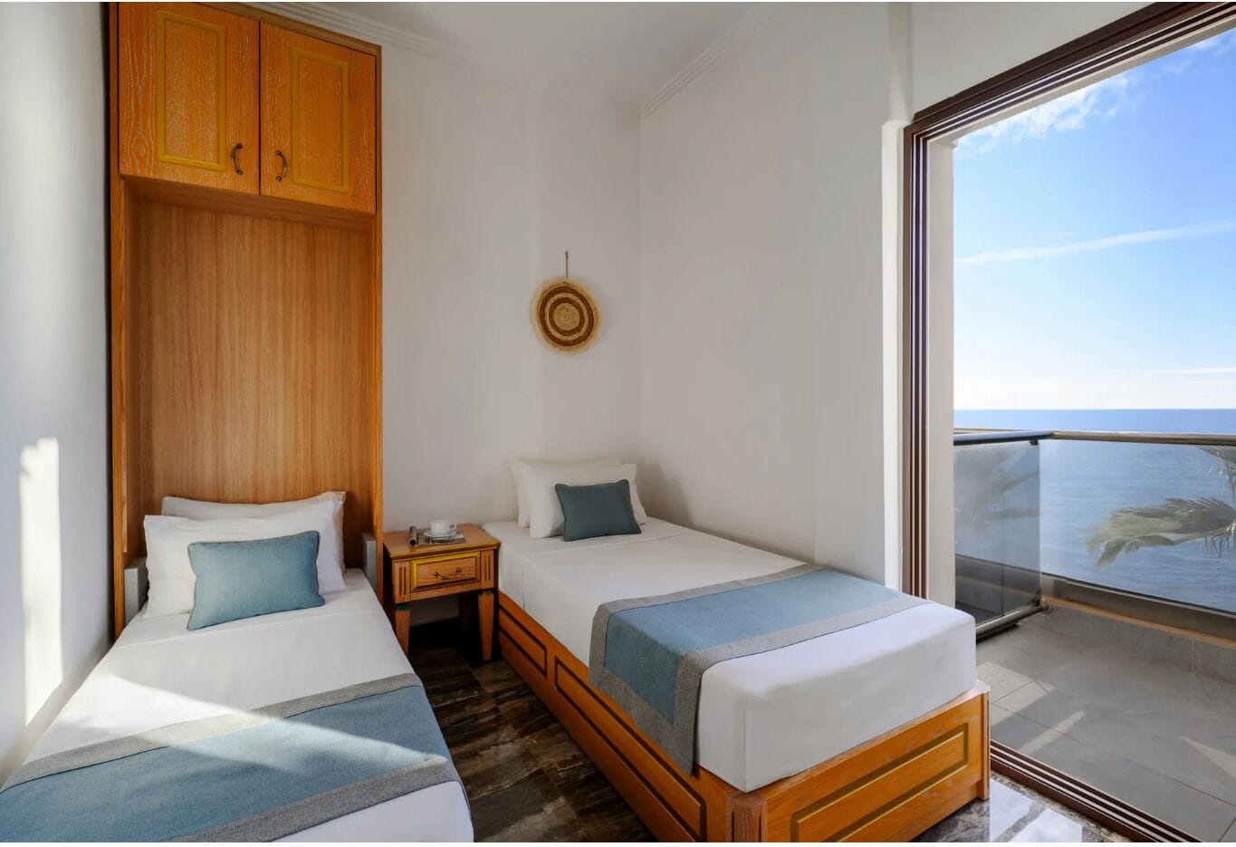 A hotel room with two beds, a wooden wardrobe, a small table with a cup, and a view of the sea through a balcony.