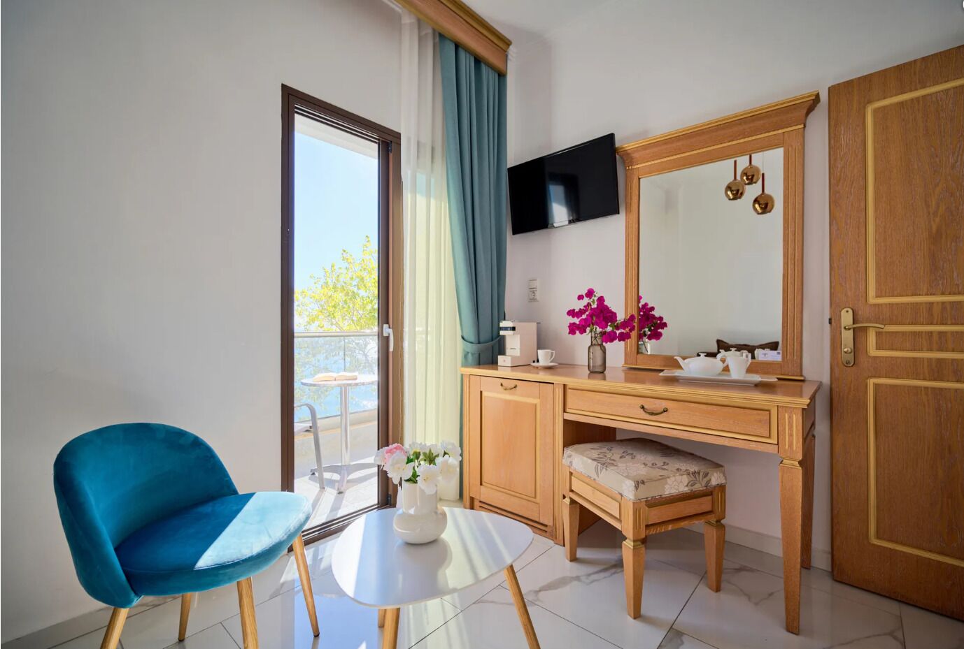Una habitación con escritorio, silla y un espejo grande. Hay un televisor montado en la pared y una puerta con pomo dorado.