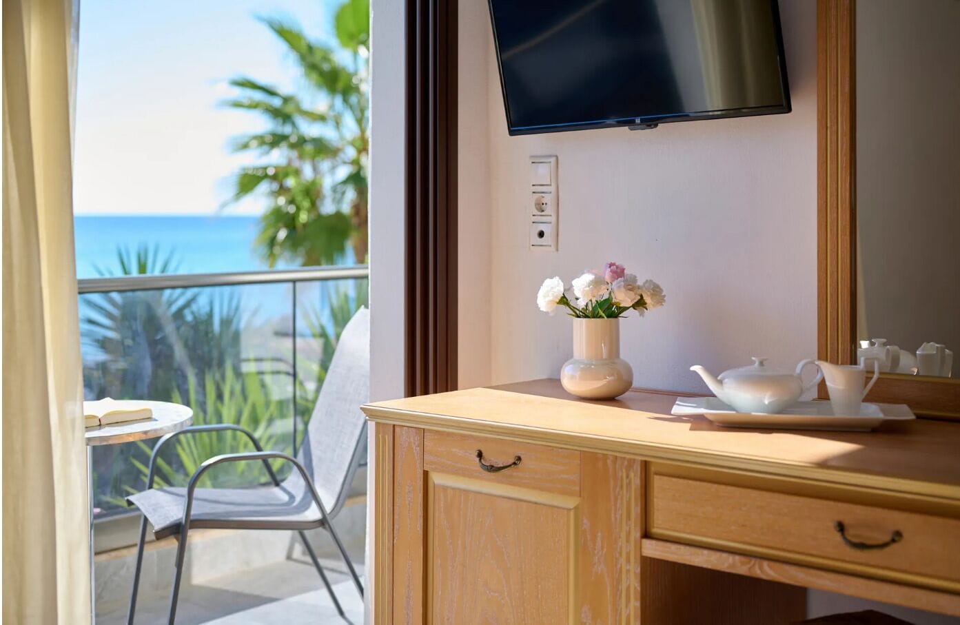 Una habitación con un escritorio de madera, un jarrón con flores, una tetera y una taza sobre una bandeja. Hay un televisor montado en la pared y una ventana con vistas al océano y palmeras.