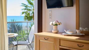 Una habitación con un escritorio de madera, un jarrón con flores, una tetera y una taza sobre una bandeja. Hay un televisor montado en la pared y una ventana con vistas al océano y palmeras.