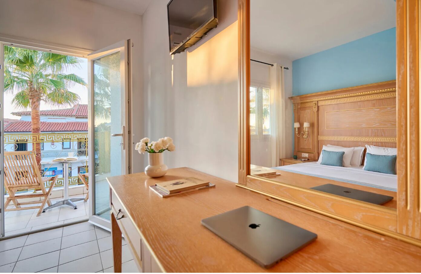 Une chambre d’hôtel avec un bureau en bois, un ordinateur portable, un vase avec des fleurs, un lit avec des oreillers et une vue sur les palmiers depuis le balcon.