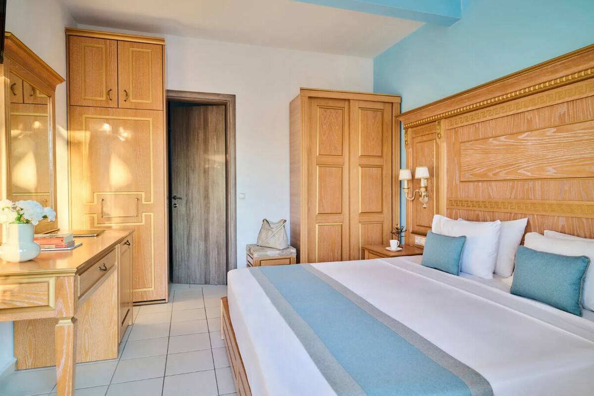 A hotel room with a large bed, wooden wardrobe, bedside table with lamps, and a small seating area.