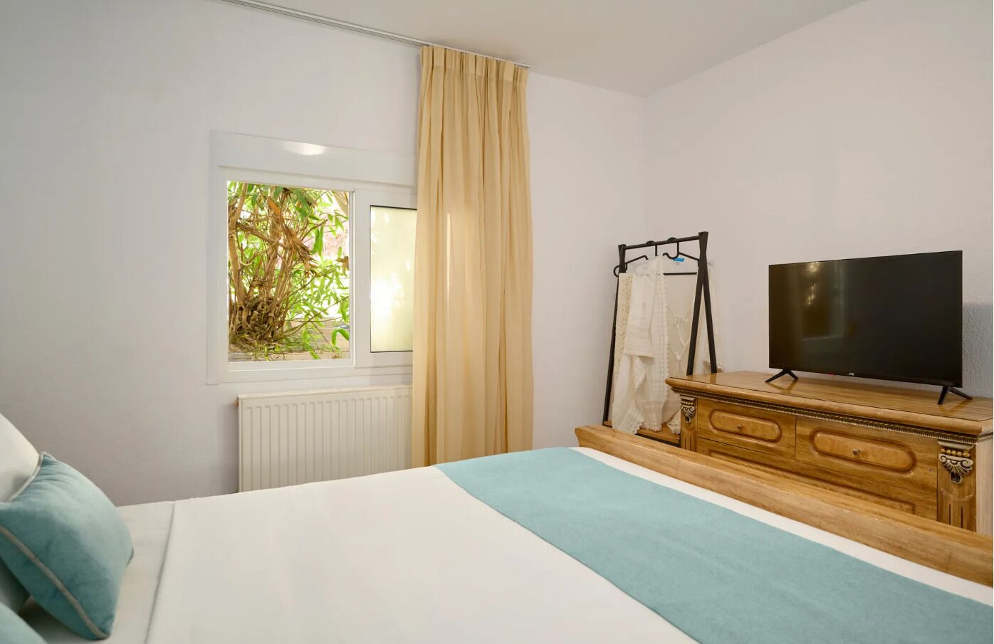 A bedroom with a bed, a TV on a wooden dresser, a window with curtains, and a radiator.