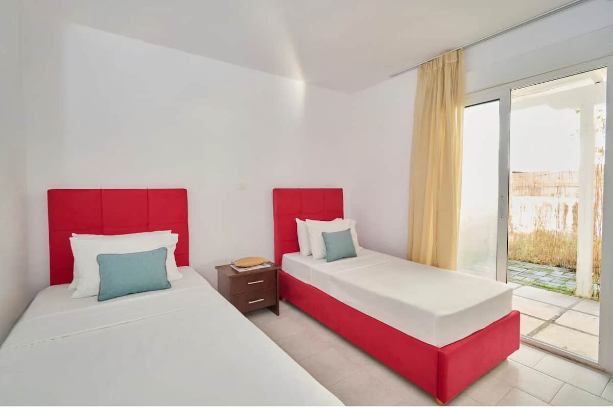 A modern hotel room with two beds, each with a red headboard and white bedding. There is a small wooden nightstand between the beds.