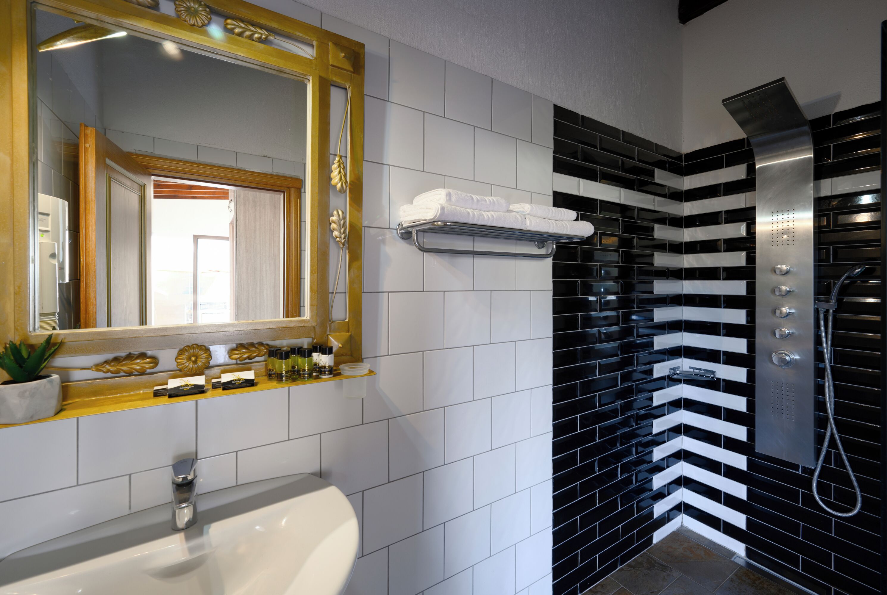 A modern bathroom with a white sink, a large mirror with a gold frame, and a black and white tiled shower area.