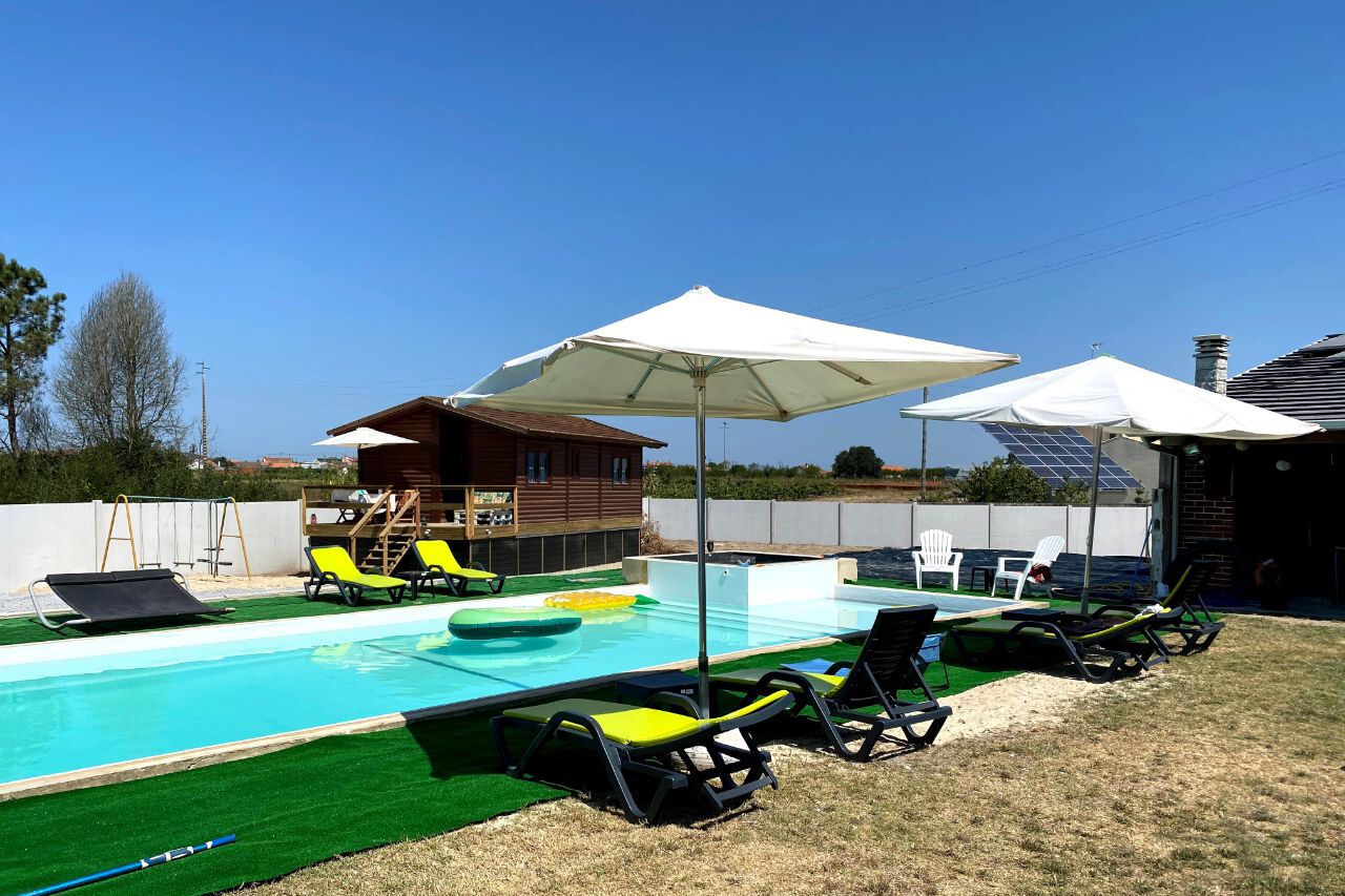 En swimmingpool med liggestole og parasoller, omgivet af en træterrasse og en mindre bygning.