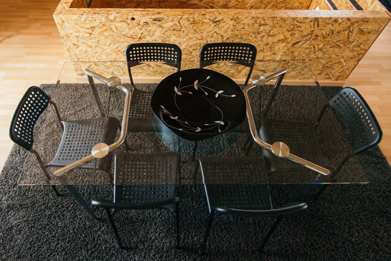Una mesa de comedor con cubierta de vidrio, cuatro sillas negras, un plato negro con diseño blanco y un aparador de madera al fondo.
