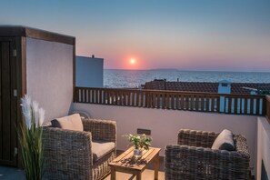 A balcony with wicker furniture, a small table, and a view of the sea at sunset.