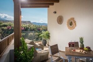 A balcony with a view of a mountainous landscape, a table with drinks and fruit, and a chair.