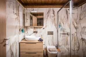 A bathroom with a marble wall, a wooden vanity, a white sink, a glass shower enclosure, and a toilet.