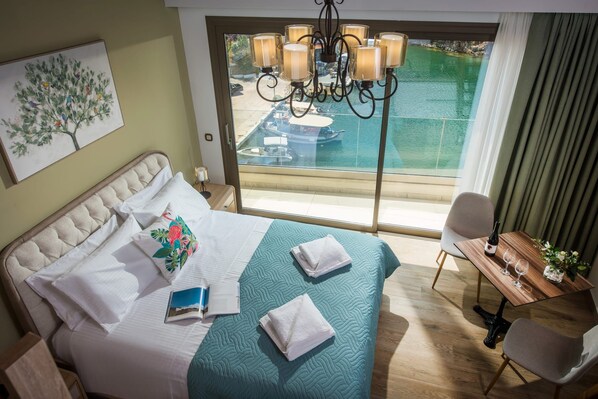 A hotel room with a bed, a dining table, a chair, a chandelier, and a view of a pool and boats.