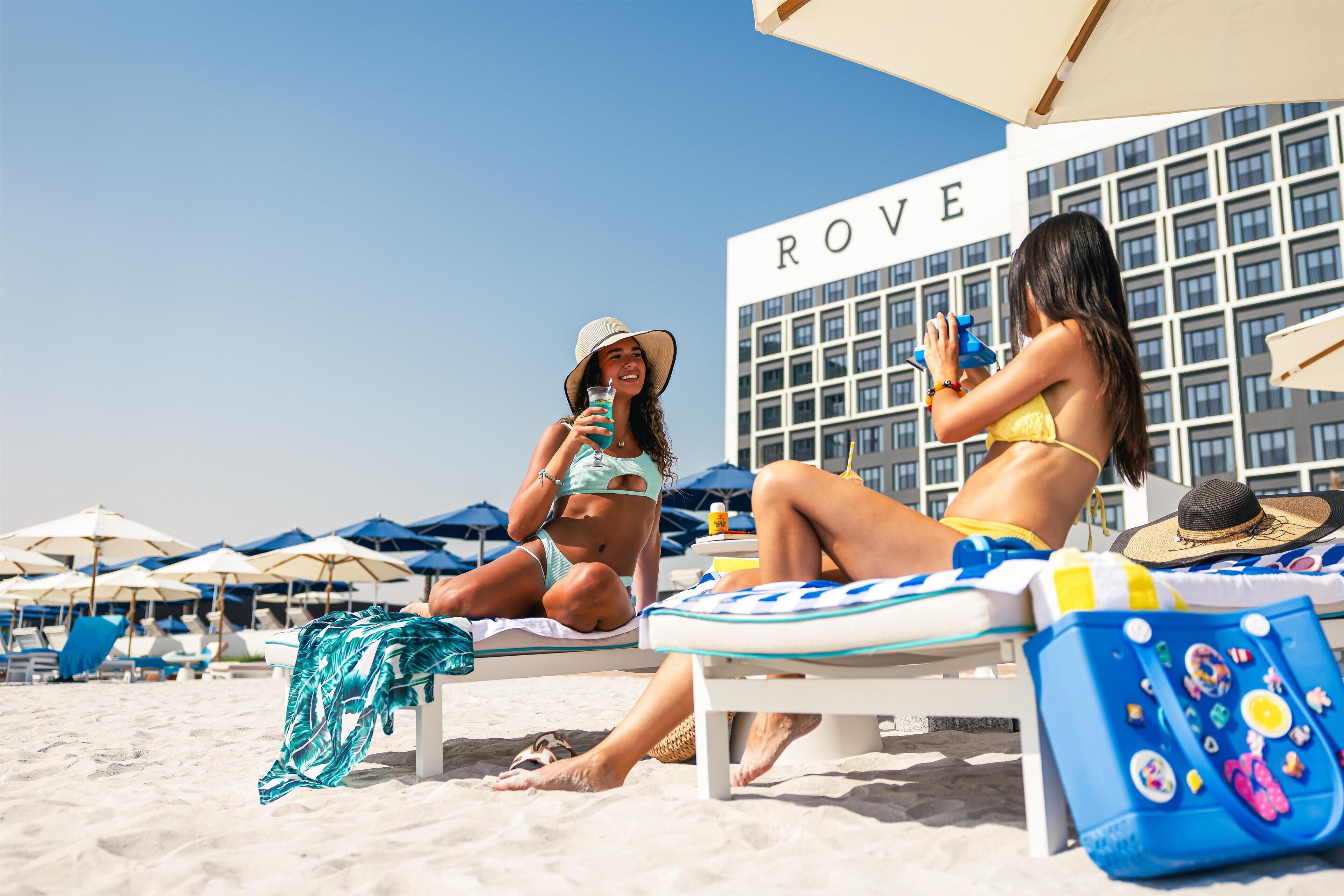 Two women relaxing on beach chairs under umbrellas at a hotel named ROVE, with a clear sky and a sandy beach in the background.