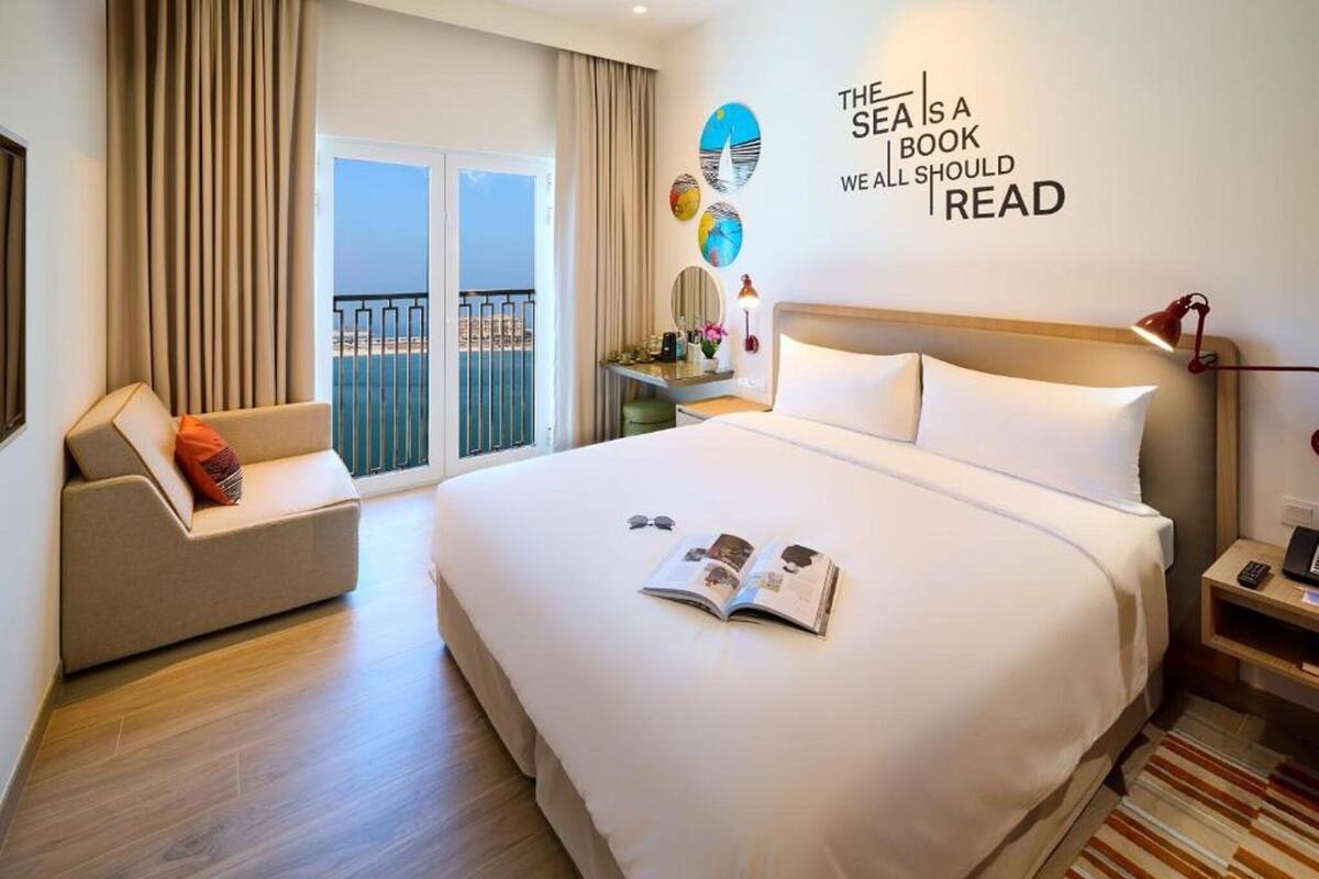Habitación de hotel con una cama grande, un sofá, un escritorio con lámpara y vista al mar.