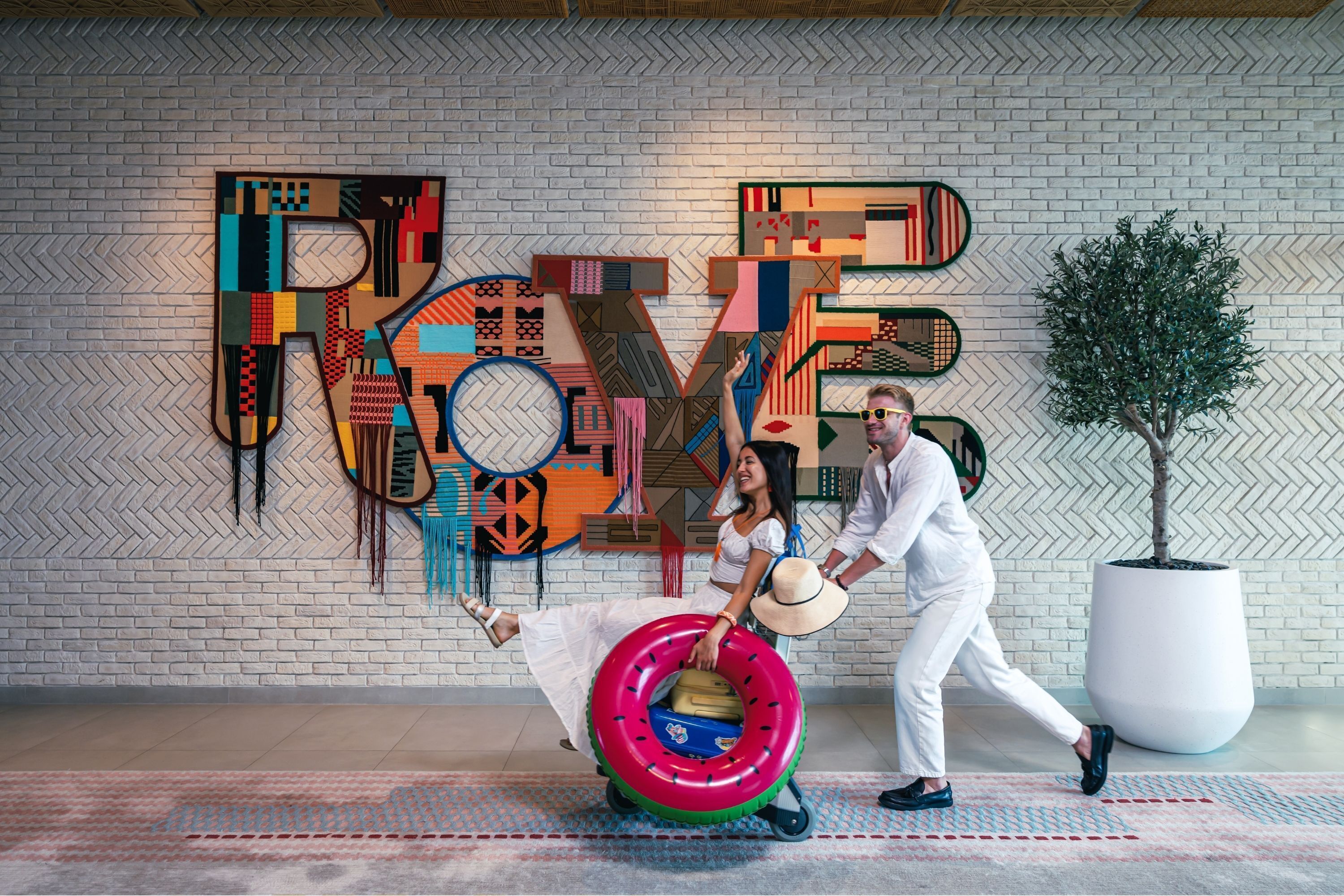 A modern interior with a large geometric 'LOVE' sign on the wall, a potted plant, and a couple posing with a suitcase.