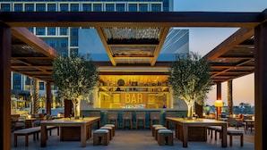 An outdoor bar area with wooden tables and chairs, a bar counter, and a view of a modern building in the background. - Rove Al Marjan Island (Ras Al Khaimah)
