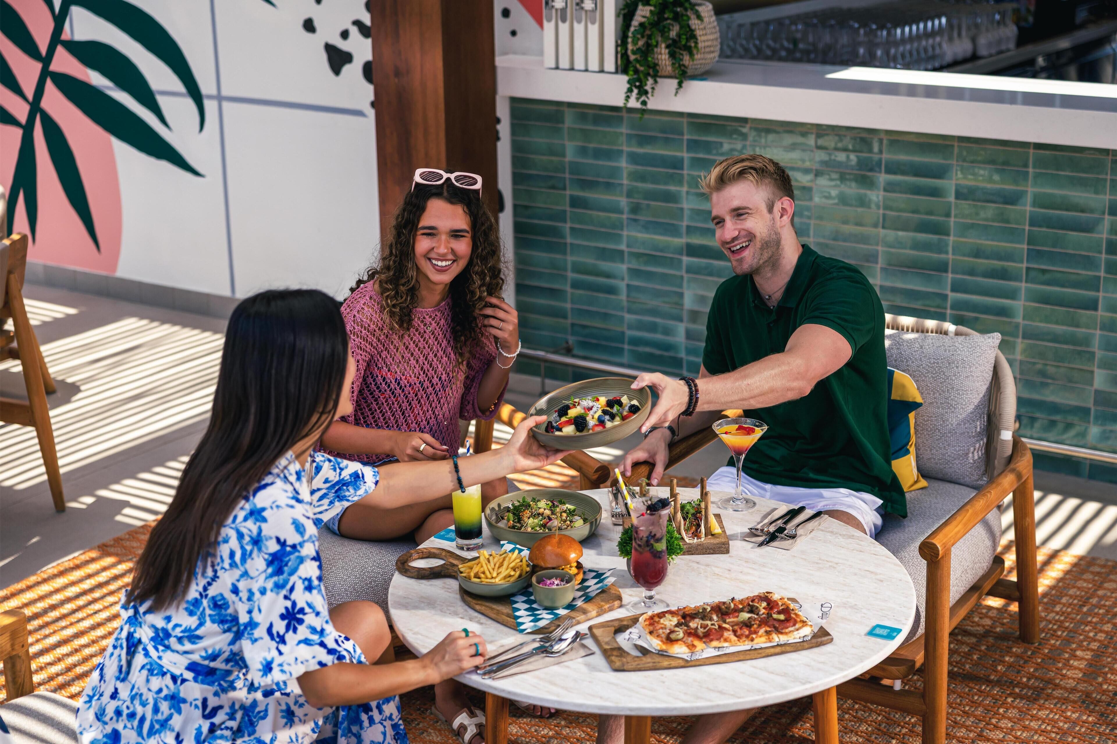 A group of people dining at an outdoor table with various dishes, drinks, and a pizza.
