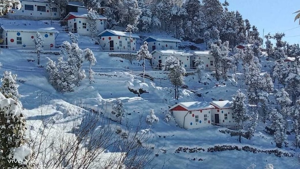 Un village enneigé, composé de plusieurs petits bâtiments, probablement des maisons d’hôtes ou des chalets, répartis sur une colline.