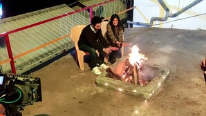 Deux personnes sont assises près d’un feu de camp dans un cadre extérieur, avec une structure en métal recouvrant le toit en arrière-plan.