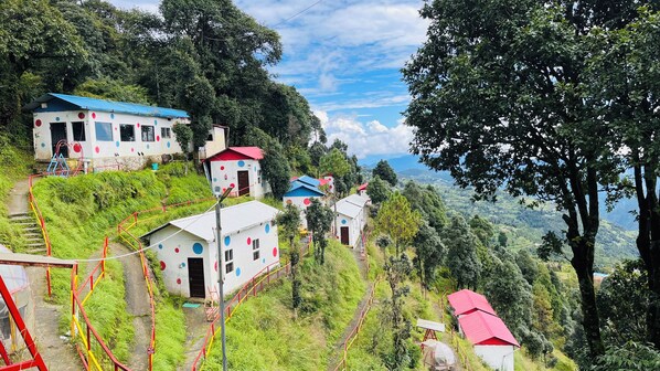A hillside with several small buildings, some with red roofs, surrounded by lush greenery and a clear sky. - Dev Bhoomi Resort (Tehri)