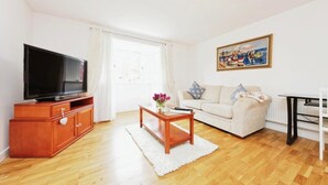 Een woonkamer met een flatscreen televisie, een houten kast, een witte bank en een schilderij aan de muur.