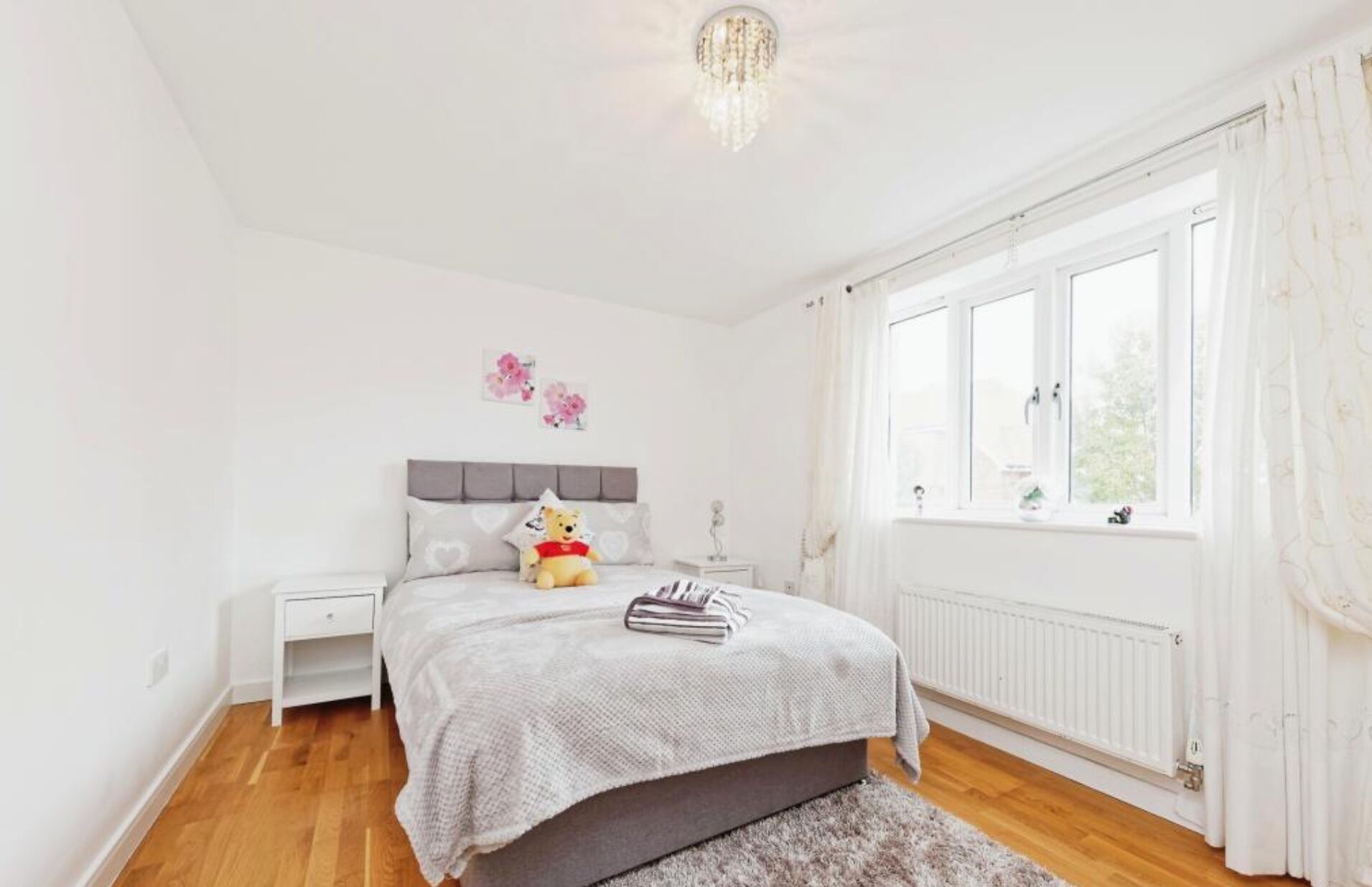 A bedroom with a bed, a nightstand, a teddy bear, a window with curtains, and a radiator.
