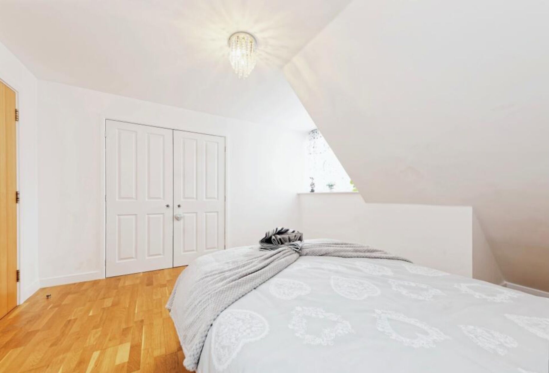 A bedroom with a wooden floor, a bed with a patterned bedspread, a white wardrobe, and a slanted ceiling with a light fixture.