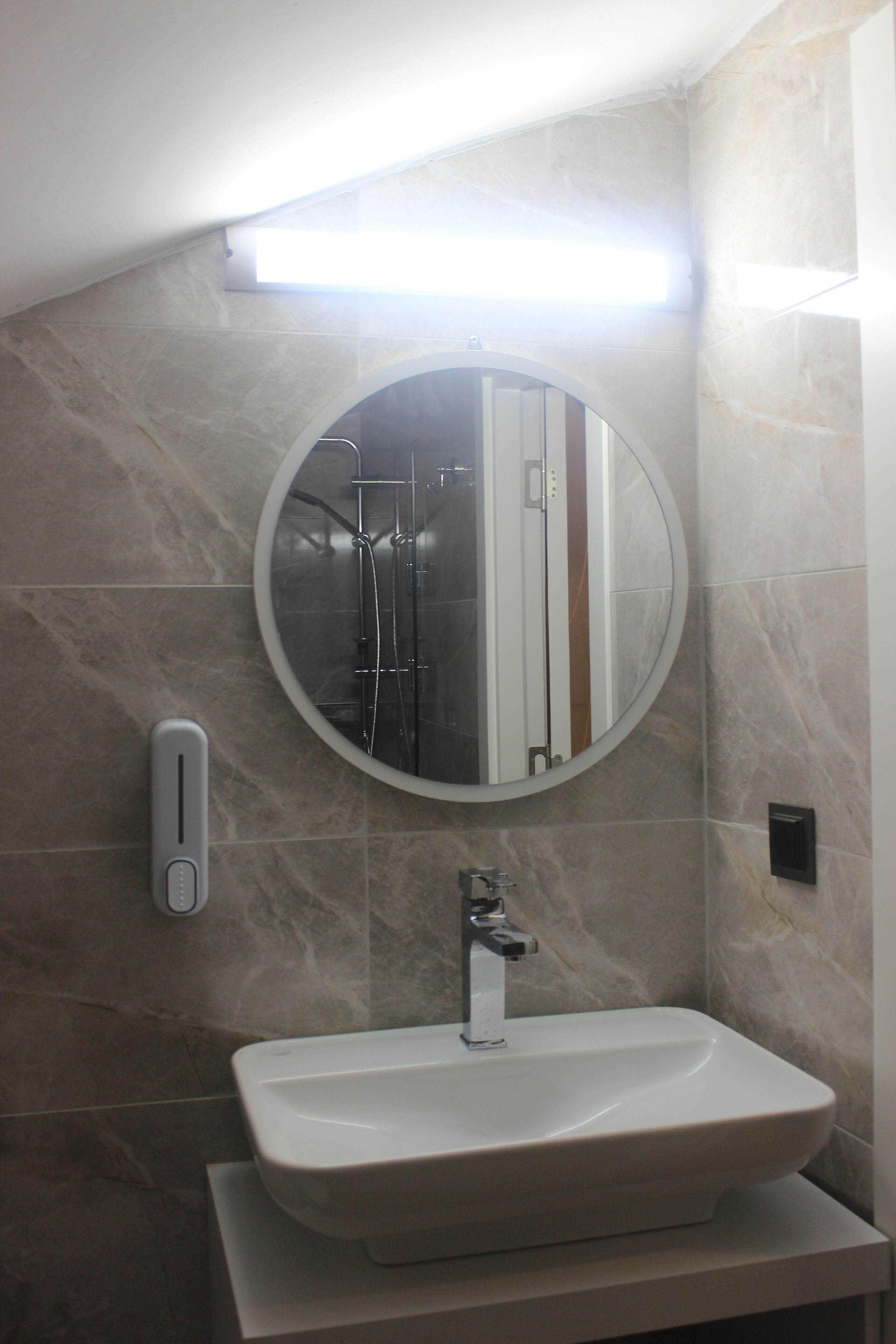 A bathroom with a round mirror, a sink, and a handheld soap dispenser.