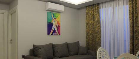 Een woonkamer met een grijze bank, witte stoelen, een eettafel en een kleurrijk poster aan de muur.