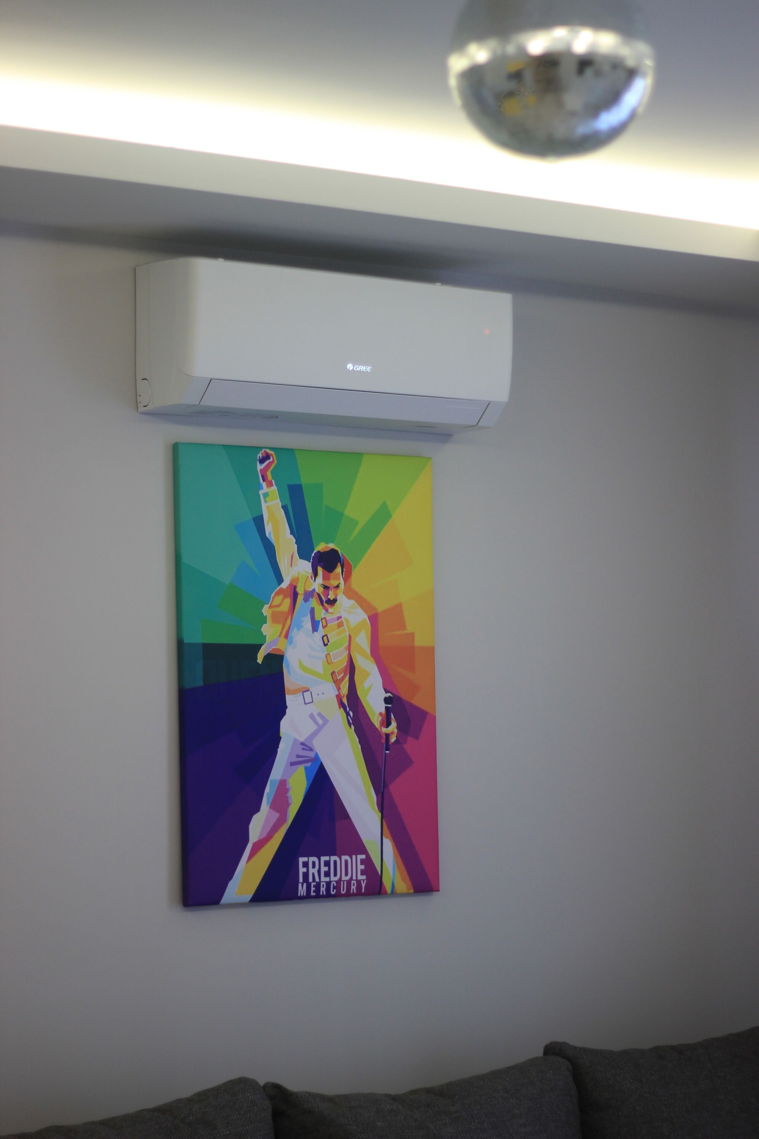 A room with a large poster of Freddie Mercury on the wall, a ceiling-mounted air conditioner, and a gray sofa.