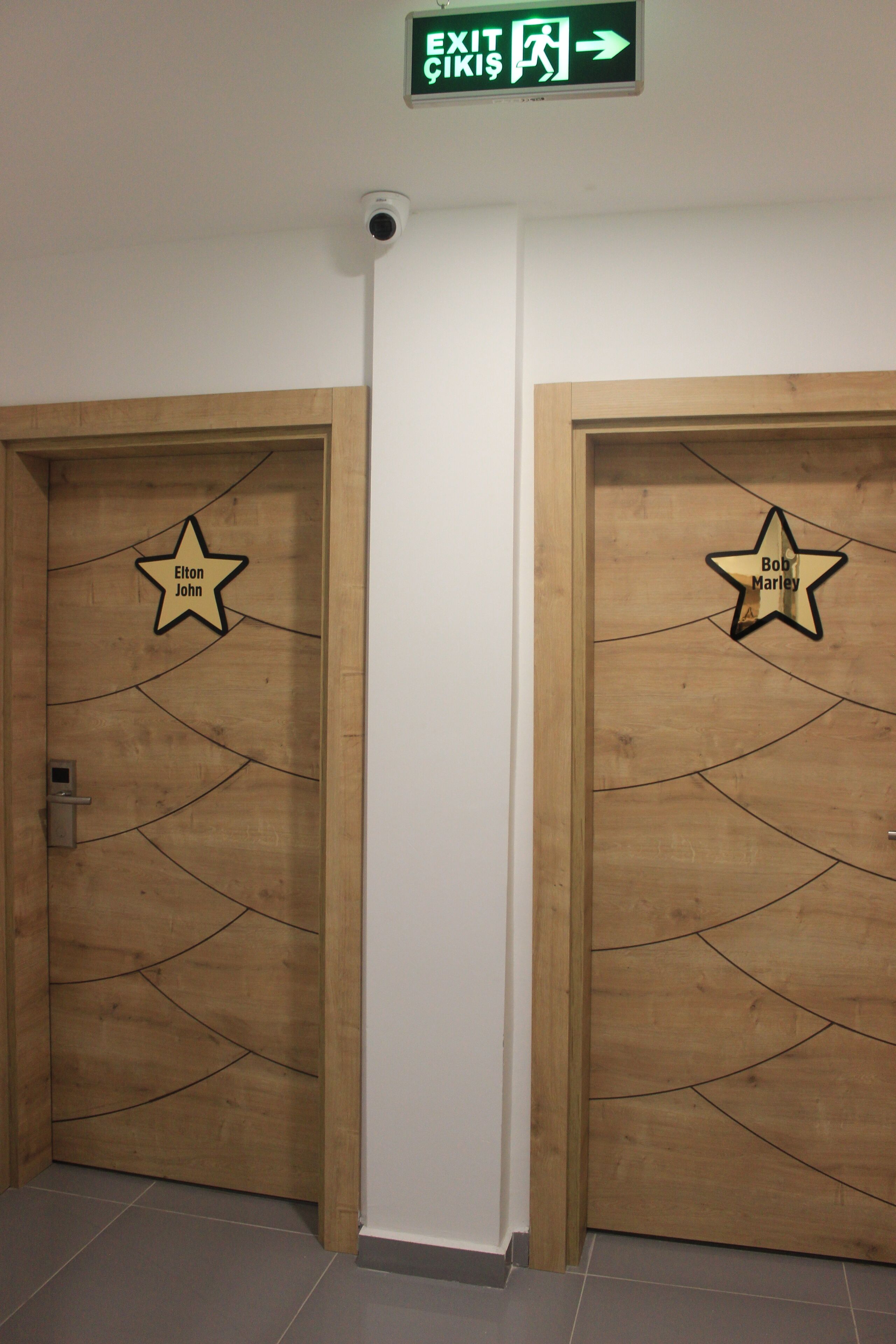 Duas portas de madeira com recortes em formato de estrela, cada uma identificada com um nome. Uma placa de saída acima das portas indica a direção da saída.