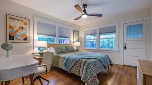 Een slaapkamer met een bed, een bureau, een stoel, een wereldbol, een plafondventilator en een raam met jaloezieën.