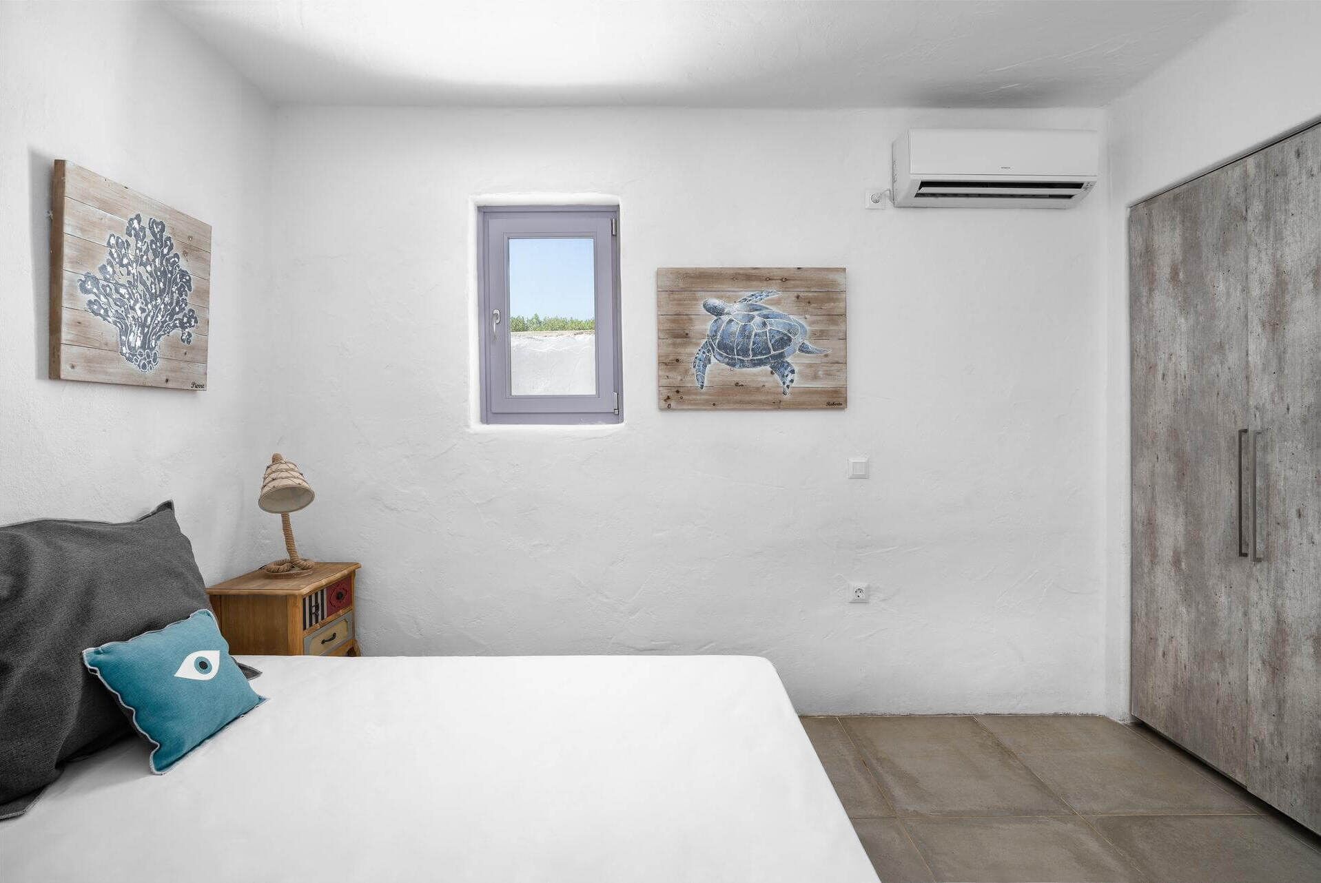 A bedroom with a bed, a nightstand, a lamp, a window, and two wall-mounted artworks.