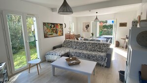 A spacious living room with a white sofa set, a coffee table, and a dining area with a table and chairs. There is a large window providing natural light and a view of greenery outside. - TYKLAN house in Bénodet (Bénodet)