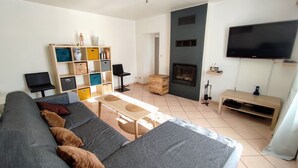 A living room with a grey sofa, a wooden coffee table, a bookshelf, a fireplace, and a TV mounted on the wall. - Comfortable cottage with fireplace and parking, near Verdun (Dompcevrin)
