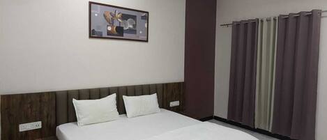 Quarto de hotel com cama, ventoinha no teto, ar condicionado fixado na parede, janela com cortinas e um quadro emoldurado na parede.
