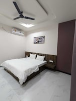 Quarto com cama, ventilador de teto, ar condicionado, um quadro pendurado na parede e uma mesa de cabeceira de madeira.