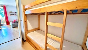 Un lit superposé avec une structure en bois et un matelas blanc, dans une pièce dotée d’un plancher en bois et d’une porte donnant sur un espace extérieur agrémenté d’un rideau rouge.