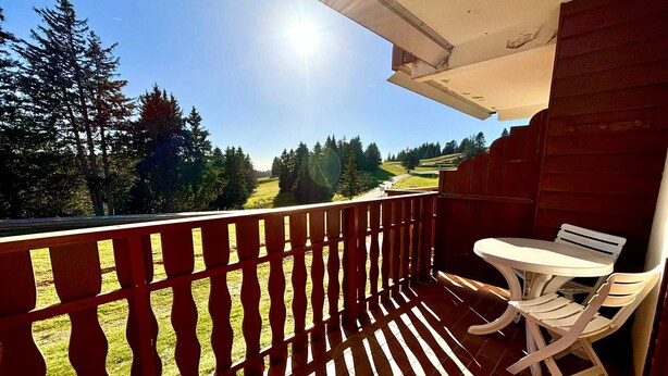 Un balcon donnant sur un paysage verdoyant et des arbres, une table blanche et deux chaises blanches.