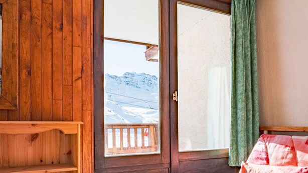 A wooden room with a bed, a window with a view of mountains, and a wooden shelf.