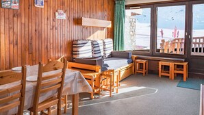 A cozy room with wooden walls, a bed with striped bedding, a dining table with chairs, and a view of a snowy landscape through large windows.