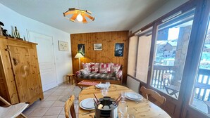 A cozy dining area with a wooden table set for four, a sofa with patterned cushions, and a view of a wooden deck through a large window. - Apartment Vars, 2 bedrooms, 4 persons (Vars)