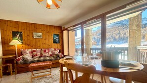 A cozy room with a wooden interior, a sofa with patterned cushions, a dining table set for two, and a view of a snowy landscape through large windows. - Apartment Vars, 2 bedrooms, 4 persons (Vars)